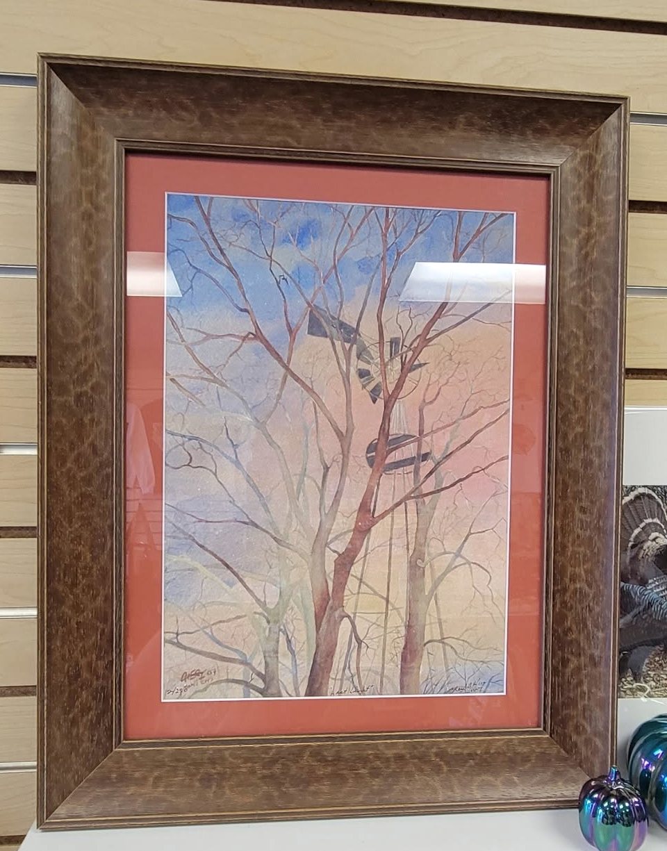 A signed watercolour of a windmill among bare trees, in a rust red mat inside a wide burl figured brown frame.